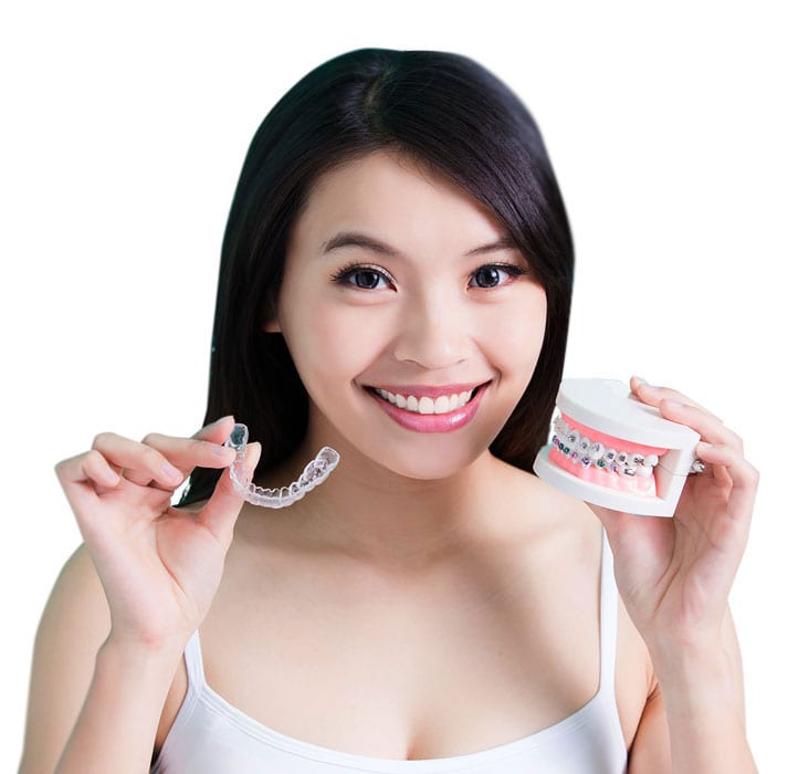 A young woman is smiling and holding a model of teeth with braces and a clear aligner in her hands.