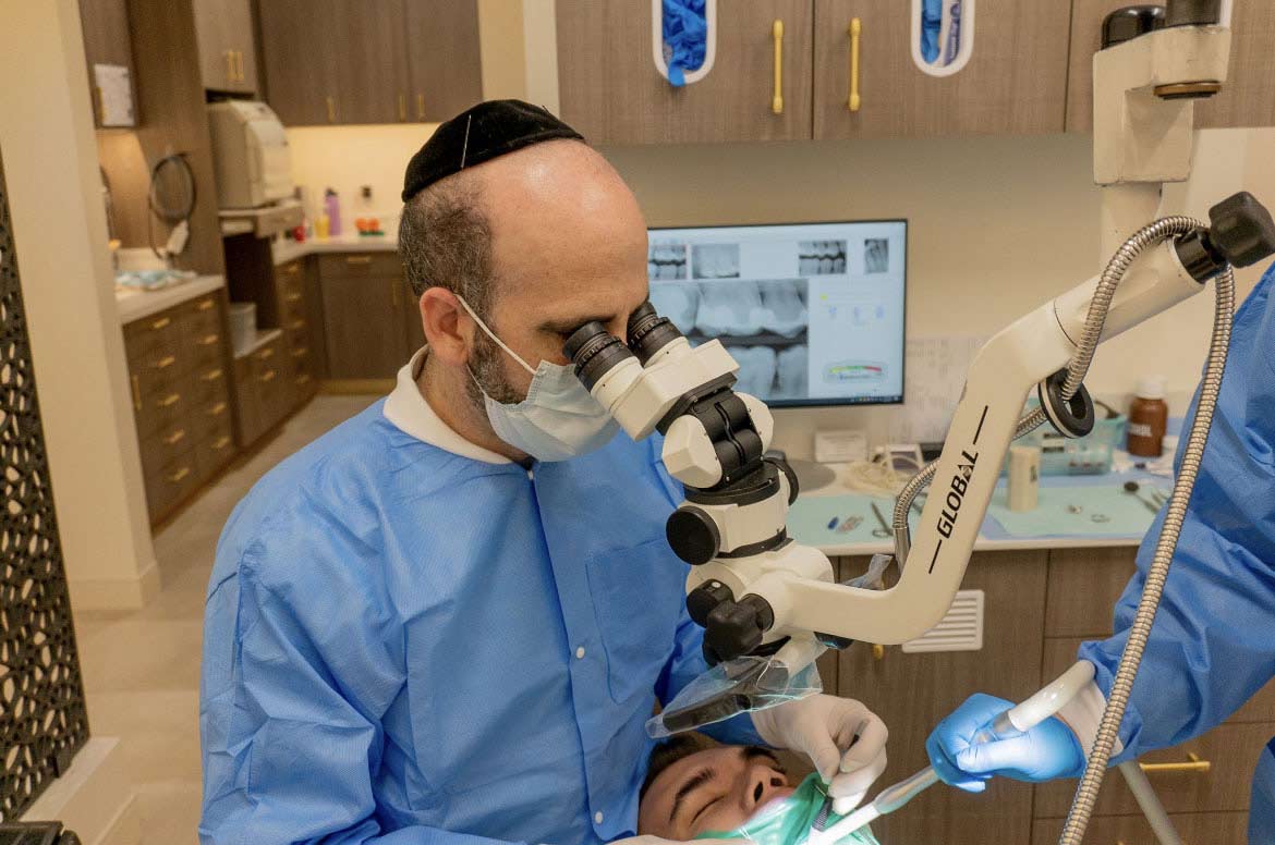 A dentist wearing a yarmulke and a blue surgical gown is performing a dental procedure on a patient while looking through a microscope.