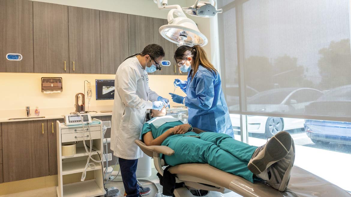 A dentist and his assistant are working on a patient's teeth. The patient is sitting in a dental chair and has their mouth open. The dentist is using a drill to work on the patient's teeth. The assistant is holding a suction tube to keep the patient's mouth dry.