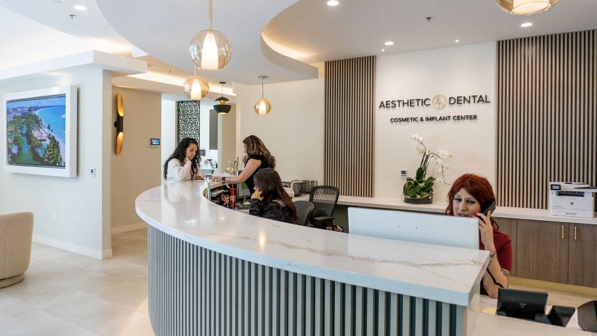 The image shows a modern dental office with a curved reception desk, wood-paneled walls, and several employees working.