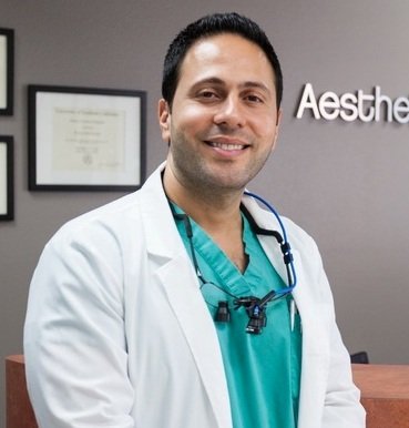 A man in a white coat and green scrubs is smiling.