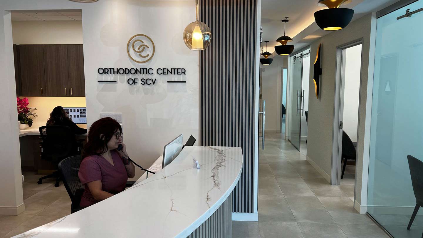 Orthodontic Center of SCV - Front Desk - The image is the inside of a modern orthodontic office. There is a woman sitting at the front desk, and the walls are decorated with wood and metal accents.