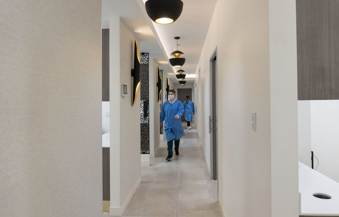 A long hallway with a tiled floor, white walls, and black light fixtures. There are two people in the hallway wearing blue scrubs and masks.