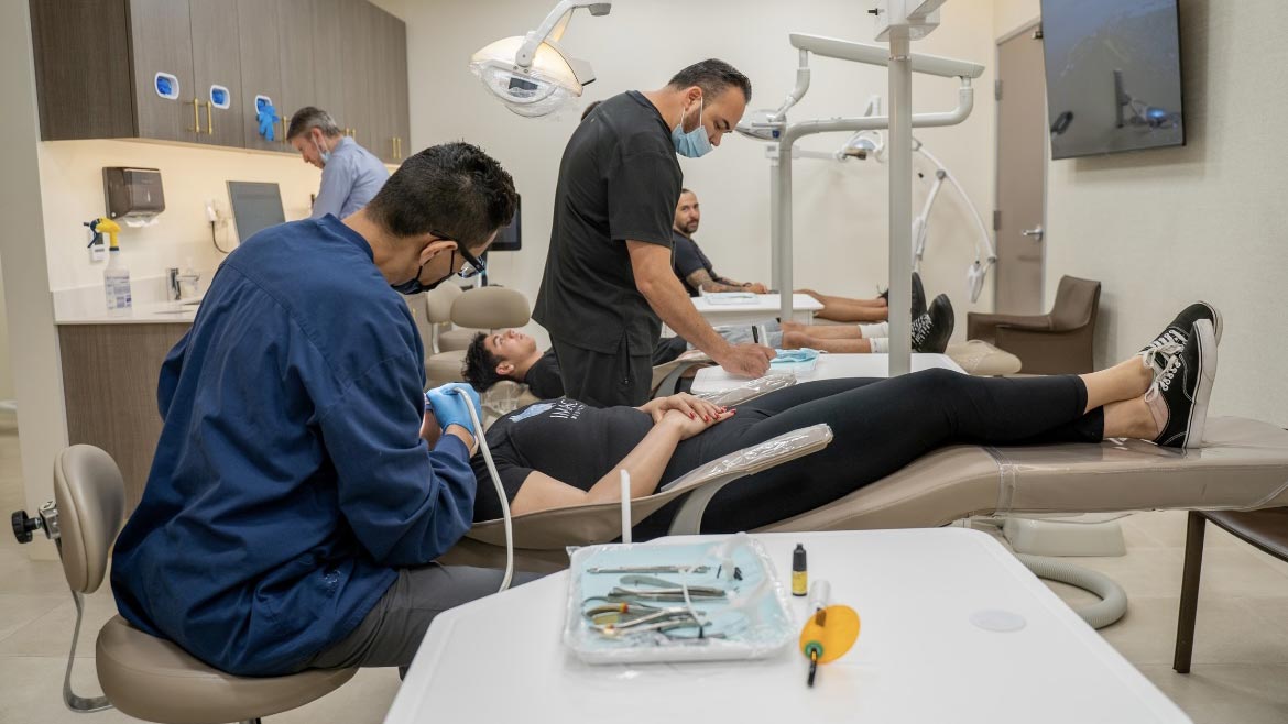 A dentist and his assistant are working on a patient's teeth. There are other patients in the background also receiving dental treatment.